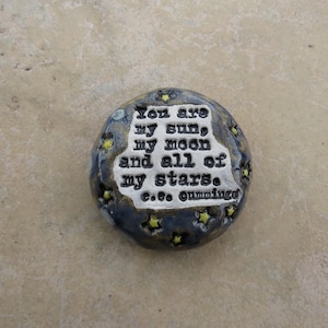 May include: A handcrafted, oval-shaped ceramic stone with a dark blue and gray speckled finish. The stone features the quote "You are my sun, my moon and all of my stars. c.e. cummings" in white lettering, with yellow star accents.