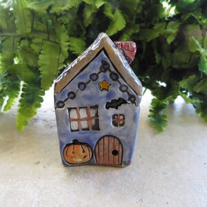 May include: A hand-painted ceramic Halloween house figurine. The blue house features a brown roof, a pumpkin, a door, and a star. A string of lights and a bat are also painted on the house. The chimney is red.