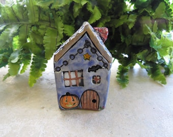 May include: A hand-painted ceramic Halloween house figurine. The blue house features a brown roof, a pumpkin, a door, and a star. A string of lights and a bat are also painted on the house. The chimney is red.