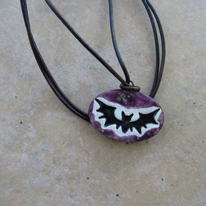 May include: A purple and white oval pendant with a black bat silhouette hanging from a brown leather cord.