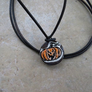 May include: A black leather cord necklace with a round pendant featuring a white and orange pumpkin with a carved jack-o'-lantern face.
