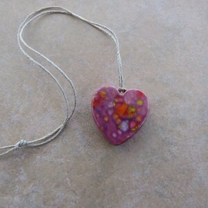 May include: A pink heart-shaped pendant with orange, yellow, and white speckles, hanging from a simple white string.