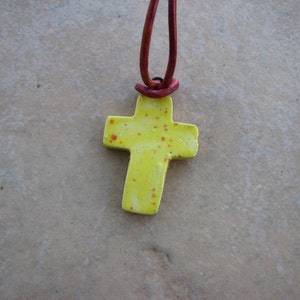 May include: A yellow ceramic cross pendant with red speckles, hanging from a red leather cord.