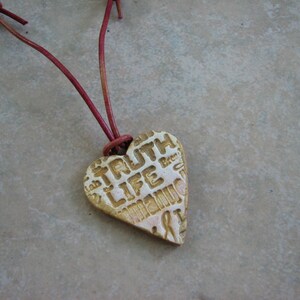 May include: A heart-shaped pendant necklace with a brown leather cord. The pendant is made of ceramic and has the words "Truth Life" embossed on it.