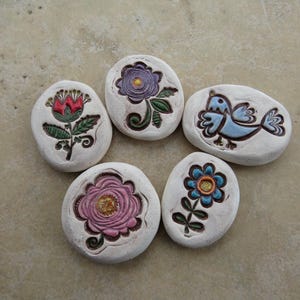 May include: A collection of five white, oval-shaped stones, each adorned with a unique hand-painted design. The designs include flowers in red, purple, and pink, and a blue bird. The stones are arranged in a circular pattern.