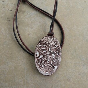 May include: Oval pendant necklace with a brown and white floral design. The pendant is suspended from a brown and black leather cord. The pendant is approximately 2 inches long.