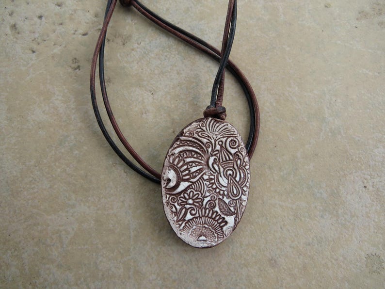 May include: Oval pendant necklace with a brown and white floral design. The pendant is suspended from a brown and black leather cord. The pendant is approximately 2 inches long.