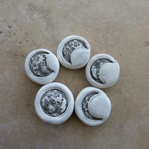 May include: Five white, circular ceramic pieces with a crescent moon design. The moon phases are in a dark gray color, and the pieces are arranged in a star-like pattern on a light brown surface.