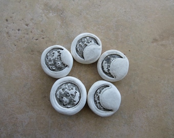 May include: Five white, circular ceramic pieces with a crescent moon design. The moon phases are in a dark gray color, and the pieces are arranged in a star-like pattern on a light brown surface.