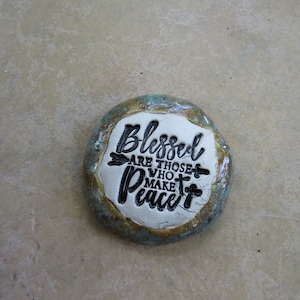 May include: A round, ceramic stone with a teal and brown glaze. The stone has a white center with the words "Blessed are those who make peace" written in black.
