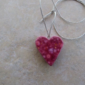 May include: A pink heart-shaped pendant with white and yellow speckles hangs from a simple white cord.