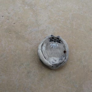 May include: A small, handmade ceramic cat-shaped dish. The dish is primarily gray with black and white details for the cat's face. The cat's tail curves around the dish. The dish is on a light brown surface.