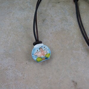 May include: A ceramic pendant necklace featuring a small, round, blue fishbowl design. Inside the fishbowl is a cartoon goldfish. The pendant hangs from a dark brown leather cord.
