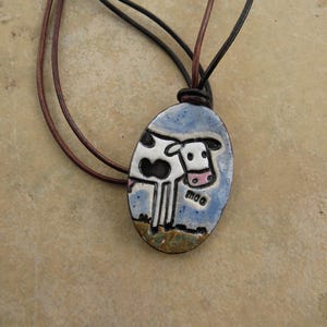 May include: Oval pendant necklace featuring a hand-painted cow design. The ceramic pendant has a blue background with a black and white cow illustration and the word "moo". The necklace has brown and black leather cords.