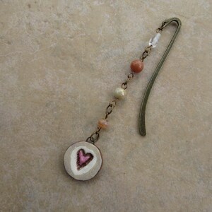 May include: A bronze bookmark with a heart-shaped charm. The charm is white with a pink heart in the center. The bookmark is decorated with small beads in shades of white, pink, and beige.
