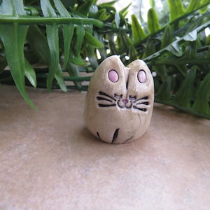 May include: A small, ceramic cat figurine with pink eyes and a white and brown painted design. The cat is sitting on a light brown surface.