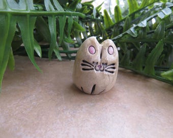 May include: A small, ceramic cat figurine with pink eyes and a white and brown painted design. The cat is sitting on a light brown surface.