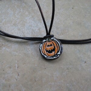 May include: A Halloween-themed necklace featuring a round pendant with a hand-painted jack-o'-lantern design. The pumpkin is orange with a black outline and a smiling face. The pendant is strung on a dark brown leather cord.