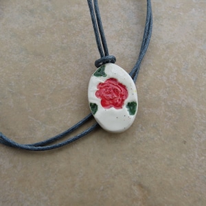 May include: A white ceramic pendant with a red rose and green leaves, hanging on a gray cord necklace.