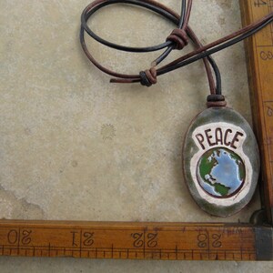 May include: Oval ceramic pendant with the word "PEACE" and a globe design in blue, green, and white. The pendant hangs from a brown and black leather cord. The background is a light beige surface.