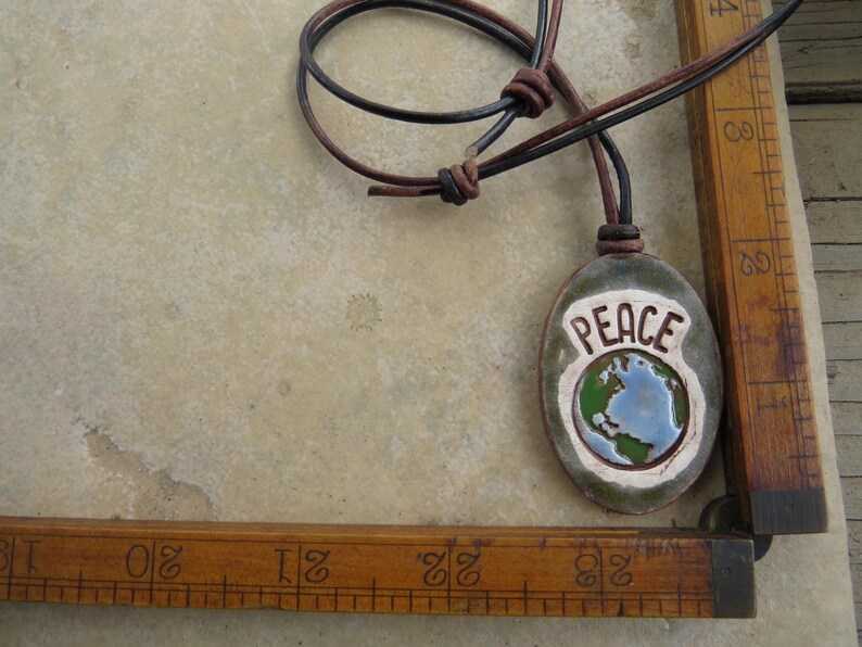 May include: Oval ceramic pendant with the word "PEACE" and a globe design in blue, green, and white. The pendant hangs from a brown and black leather cord. The background is a light beige surface.