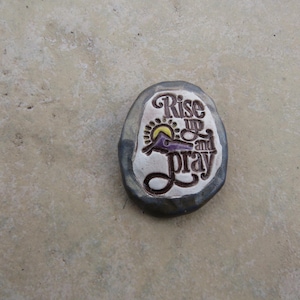 May include: Oval-shaped stone with the words "Rise up and Pray" in a decorative font. The design includes a sun and a figure in purple. The stone has a silver border and is set against a neutral background.