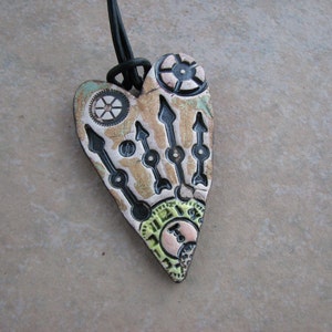 May include: A heart-shaped pendant necklace with a black cord. The pendant is made of ceramic and features a black, white, and green design with gears and arrows.