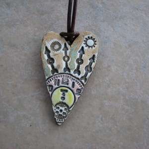 May include: A heart-shaped ceramic pendant with a clock face, arrows, and sun designs. The pendant is painted in shades of brown, white, and yellow.