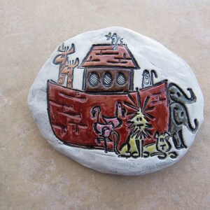 May include: A ceramic tile depicting a red ark with a brown roof and various animals, including a giraffe, a lion, a flamingo, and an elephant. The tile is painted in a whimsical style with black outlines.
