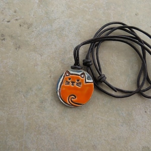 May include: A round ceramic pendant featuring a bright orange cat with black outlines, suspended on a dark brown leather cord. The cat design includes pointed ears, whiskers, and a curled tail, set against a light gray background on the pendant.