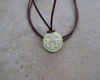 May include: A brown leather cord necklace with a round pendant featuring two white faces carved in relief. The pendant is a light yellow color.