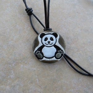 May include: A black leather cord necklace with a round pendant featuring a white panda bear with black eyes and ears. The pendant has a brown background.