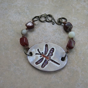 May include: A brown and white ceramic bracelet with a dragonfly design. The bracelet is made with brown cord and has red and white beads.