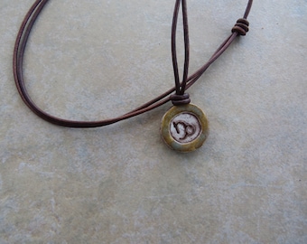 May include: A brown leather cord necklace with a circular pendant. The pendant has a white center with the Capricorn zodiac symbol and a gold-colored border. The necklace is adjustable.