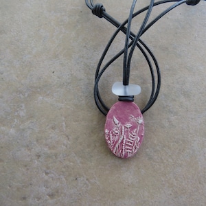 May include: A pink oval ceramic pendant with a white floral design hangs from a black leather cord. The pendant is attached to the cord with a clear glass bead.