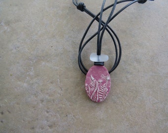 May include: A pink oval ceramic pendant with a white floral design hangs from a black leather cord. The pendant is attached to the cord with a clear glass bead.