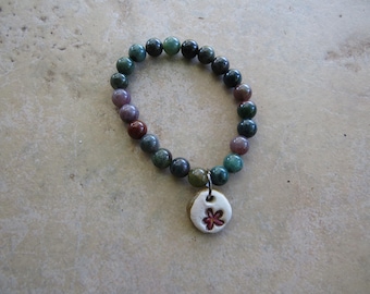May include: A beaded bracelet with a variety of colors, including green, purple, and red, is displayed. The bracelet has a small, round, white charm with a red flower design. The beads are round and appear to be made of stone.