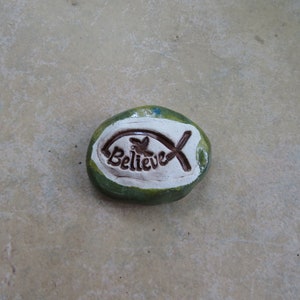 May include: A green and white ceramic stone shaped like a fish with the word "Believe" inscribed inside.