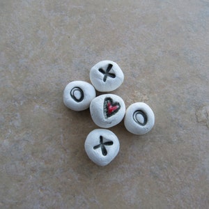 May include: Five white round stones with different symbols painted on them. The symbols include a heart, a cross, a circle, and two ovals.