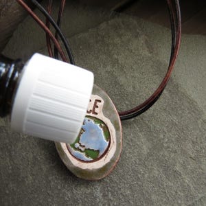 May include: A brown glass bottle with a white cap is positioned near a ceramic pendant. The pendant features a painted Earth design with the word "PEACE" and is strung on a leather cord. The pendant is oval-shaped with a brown border.