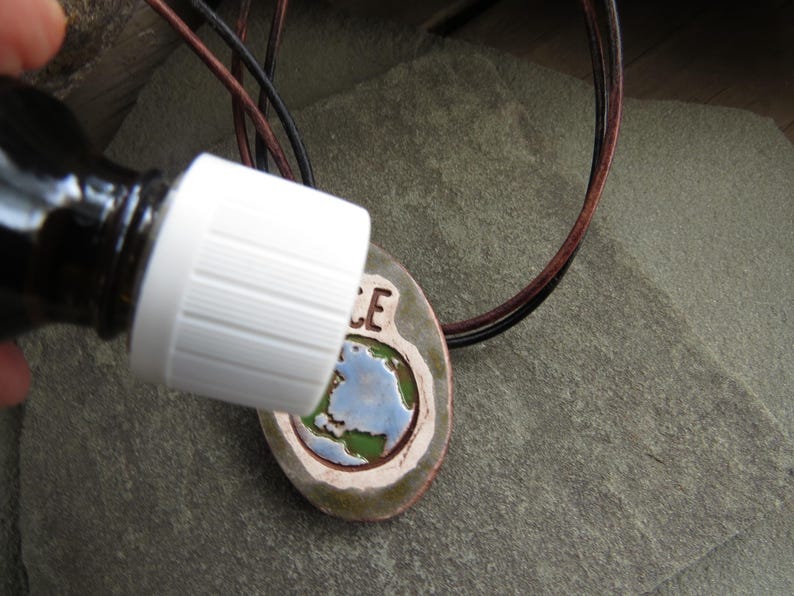 May include: A brown glass bottle with a white cap is positioned near a ceramic pendant. The pendant features a painted Earth design with the word "PEACE" and is strung on a leather cord. The pendant is oval-shaped with a brown border.