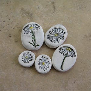 May include: A collection of six white, smooth stones, each hand-painted with a detailed daisy design. The daisies feature black outlines, yellow centers, and green stems. The stones vary in size and shape, creating a natural, artistic display.