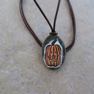 May include: A brown leather cord necklace with a ceramic pumpkin pendant. The pumpkin is orange with a white face and green stem.