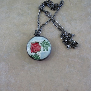 May include: A silver chain necklace with a round pendant featuring a red rose with green leaves on a white background.