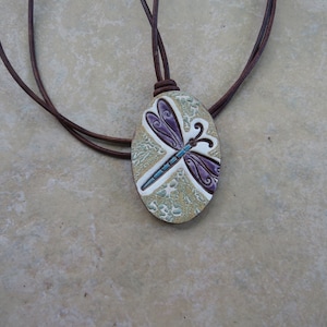 May include: A brown leather cord necklace with an oval pendant featuring a purple and blue dragonfly design on a white background.
