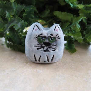May include: A small, gray ceramic cat figurine with a grumpy expression. The cat has green eyes, black whiskers, and a black nose. The ears have pink accents. The figurine is sitting in front of green foliage.