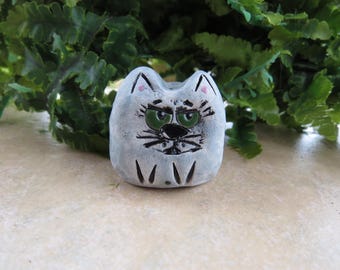 May include: A small, gray ceramic cat figurine with a grumpy expression. The cat has green eyes, black whiskers, and a black nose. The ears have pink accents. The figurine is sitting in front of green foliage.