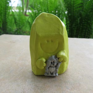 May include: A yellow ceramic figurine of a person holding a small gray dog. The person has a smiling face and is wearing a hooded robe.