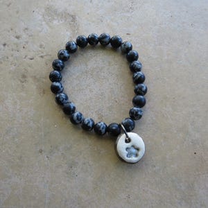 May include: A beaded bracelet featuring dark gray and white speckled beads. A small, round, silver-colored charm with a star cutout hangs from the bracelet. The bracelet is displayed on a light brown surface.