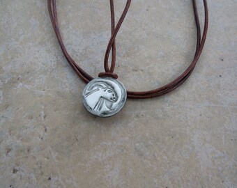 May include: A silver pendant necklace with a detailed image of a goat's head on a round disc. The necklace is on a brown leather cord.